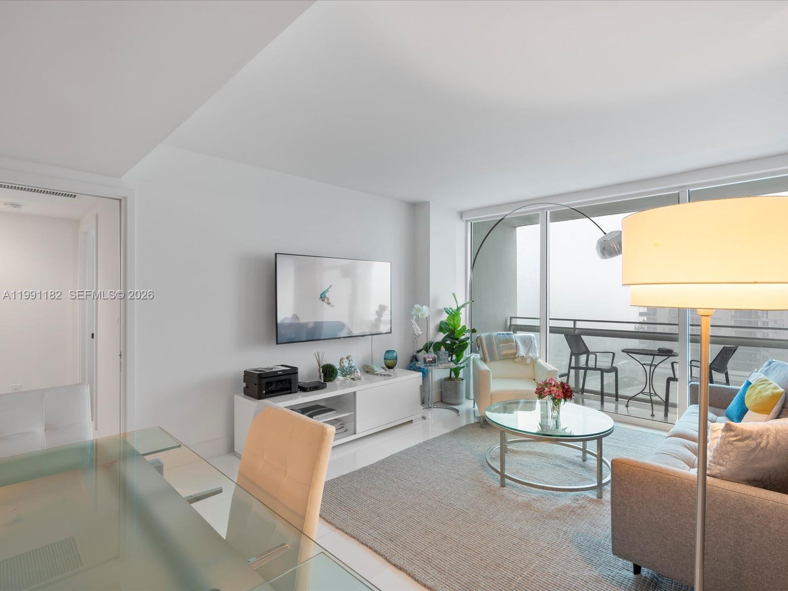 A modern living room features white flooring, a wall-mounted television, and a balcony with seating — 6801 Collins Ave # 1208, Miami Beach FL 33141, MIAMI BEACH, FL
