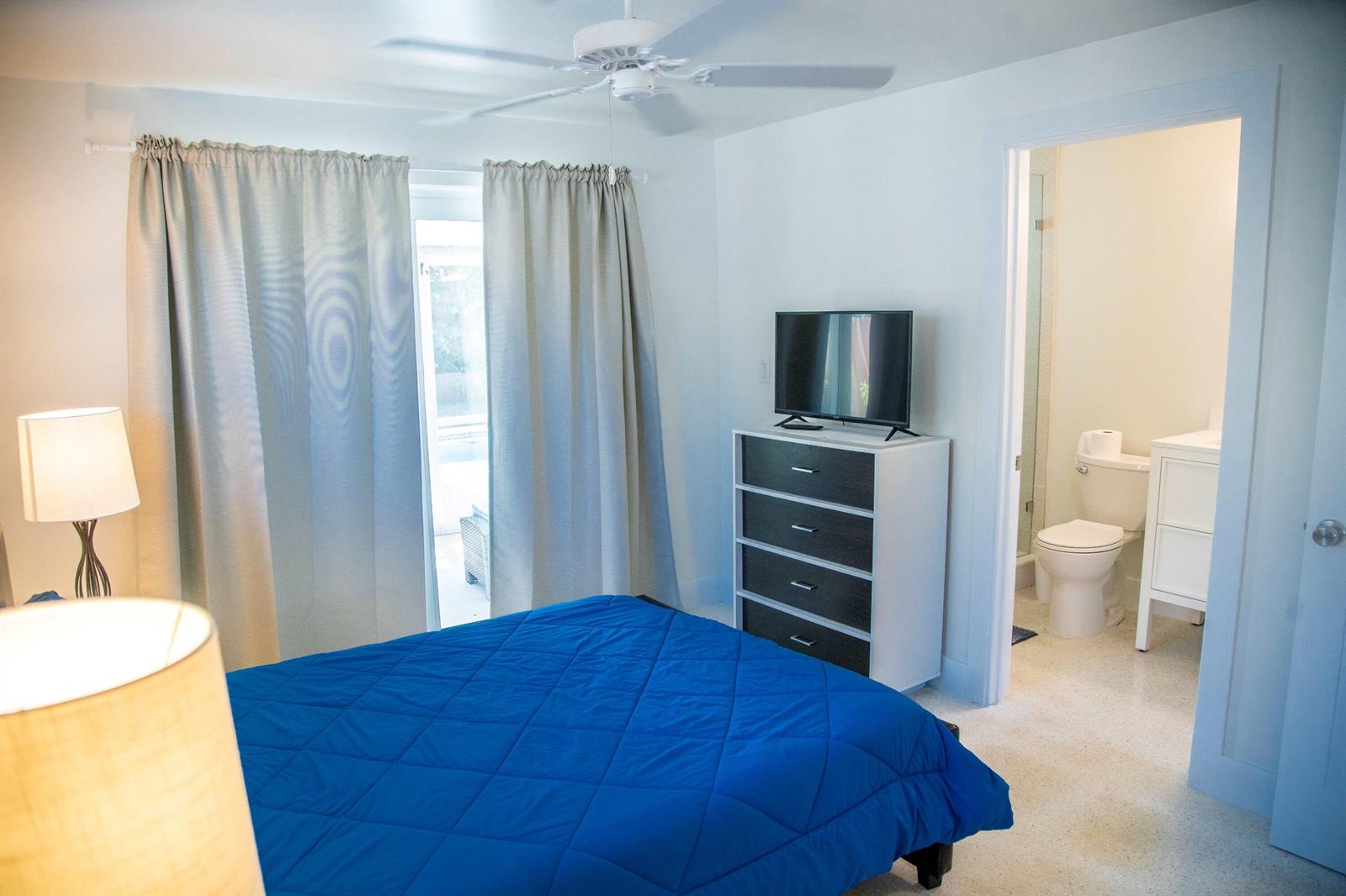 A bedroom with a blue bed, a dresser with a television, and an attached bathroom — 22 Van Road, Jupiter FL 33469, JUPITER, FL