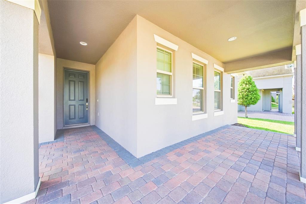 A covered entryway features a gray front door, beige stucco walls, and brick paver flooring — 10174 MERRYMEETING BAY DRIVE, WINTER GARDEN, FL