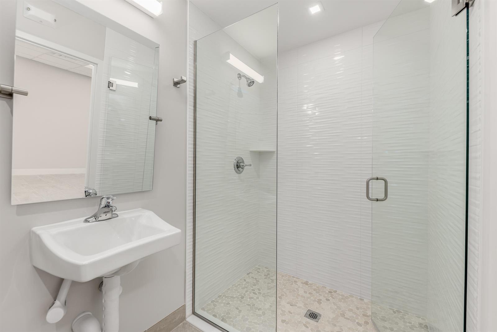 A modern bathroom features a white wall-mounted sink, a glass-enclosed shower, and pebble-tiled flooring — 41 SW Seminole Street # 210, Stuart FL 34994, STUART, FL