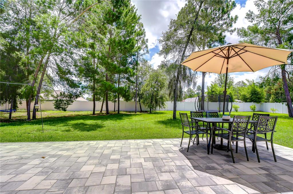 A paved patio with a dining set and umbrella overlooks a grassy, tree-lined backyard — 3358 PONYTAIL COURT, SPRING HILL, FL