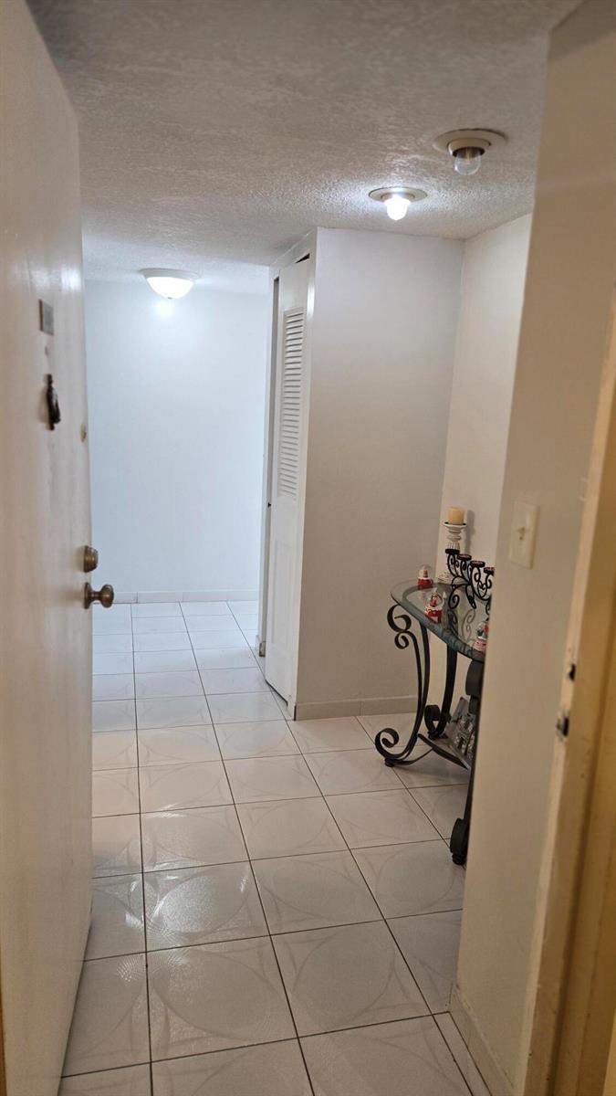 A hallway with white tile flooring leads to a white door and a decorative console table — 4164 Inverrary Drive # 601, Lauderhill FL 33319, LAUDERHILL, FL