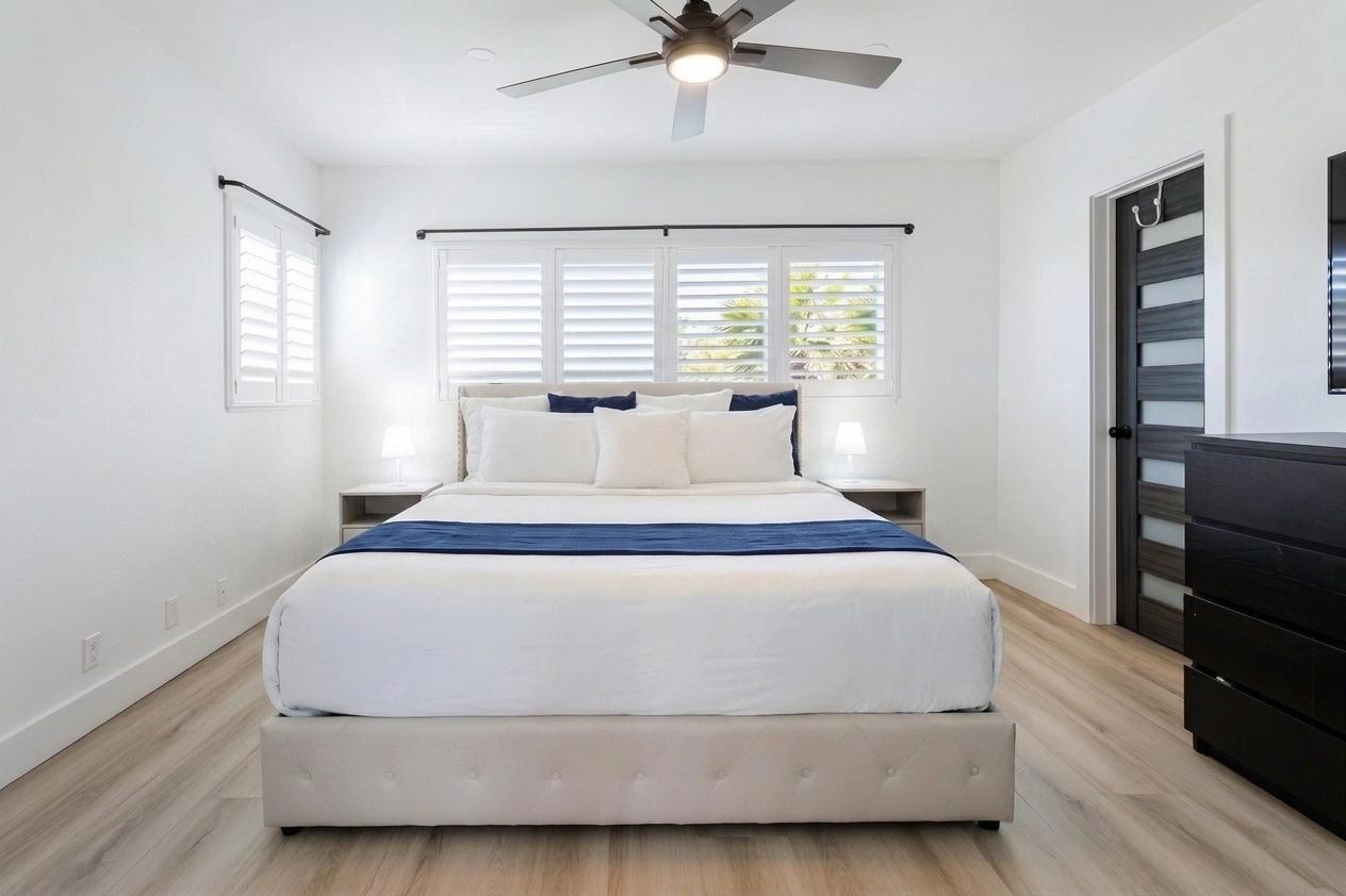 A bedroom features a tufted bed, white bedding, plantation shutters, and light wood-look flooring — 1201 S Riverside Drive # 208, Pompano Beach FL 33062, POMPANO BEACH, FL