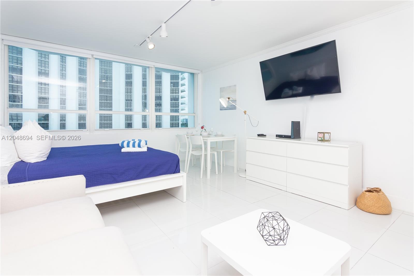 A bright studio apartment features white tile floors, a bed, dining set, and wall-mounted television — 5445 Collins Ave # 908, Miami Beach FL 33140, MIAMI BEACH, FL