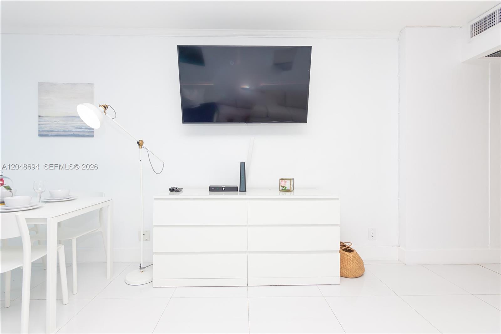 A white room features a wall-mounted television above a white dresser and dining table — 5445 Collins Ave # 908, Miami Beach FL 33140, MIAMI BEACH, FL