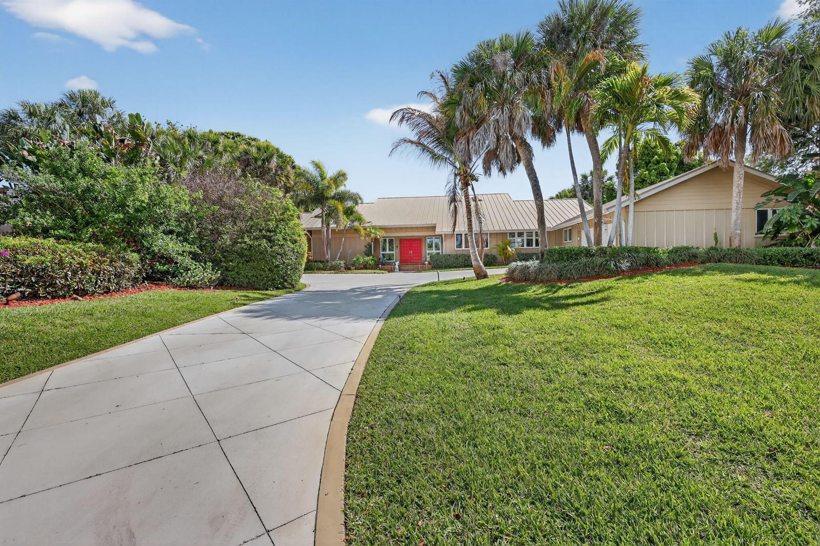 A tan house with a red front door and a concrete driveway sits among palm trees — 6151 SE Winged Foot Drive, Stuart FL 34997, STUART, FL