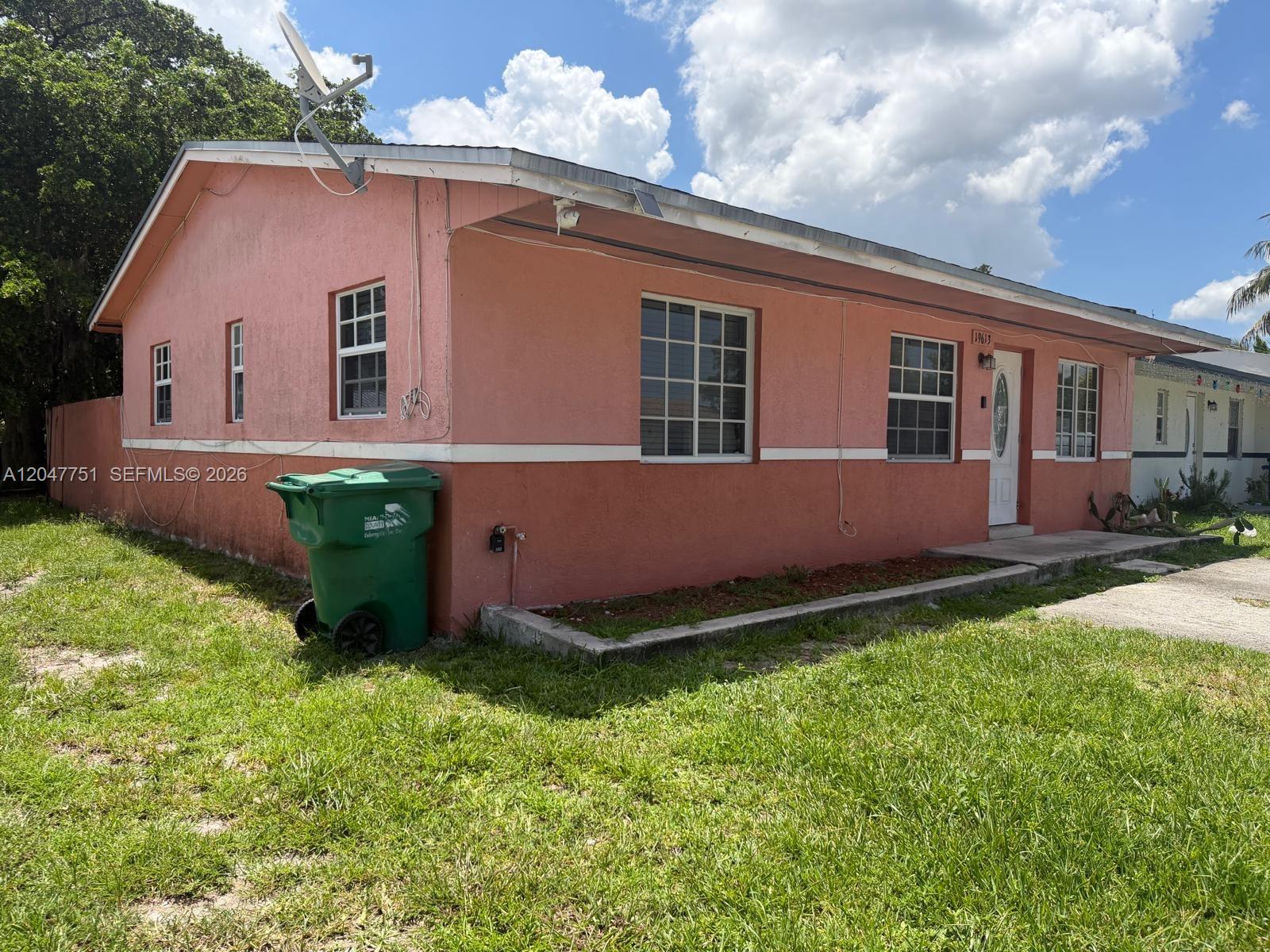 A single-story pink house with a green trash bin sits on a grassy lawn — MIAMI GARDENS, FL
