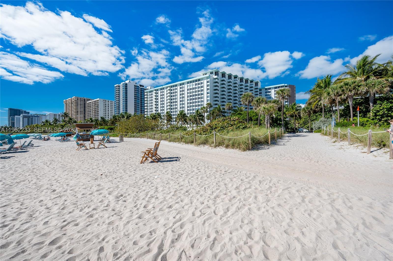 A sandy beach path leads toward large residential buildings under a bright blue sky — 2899 Collins Ave # 1505, Miami Beach FL 33140, MIAMI BEACH, FL