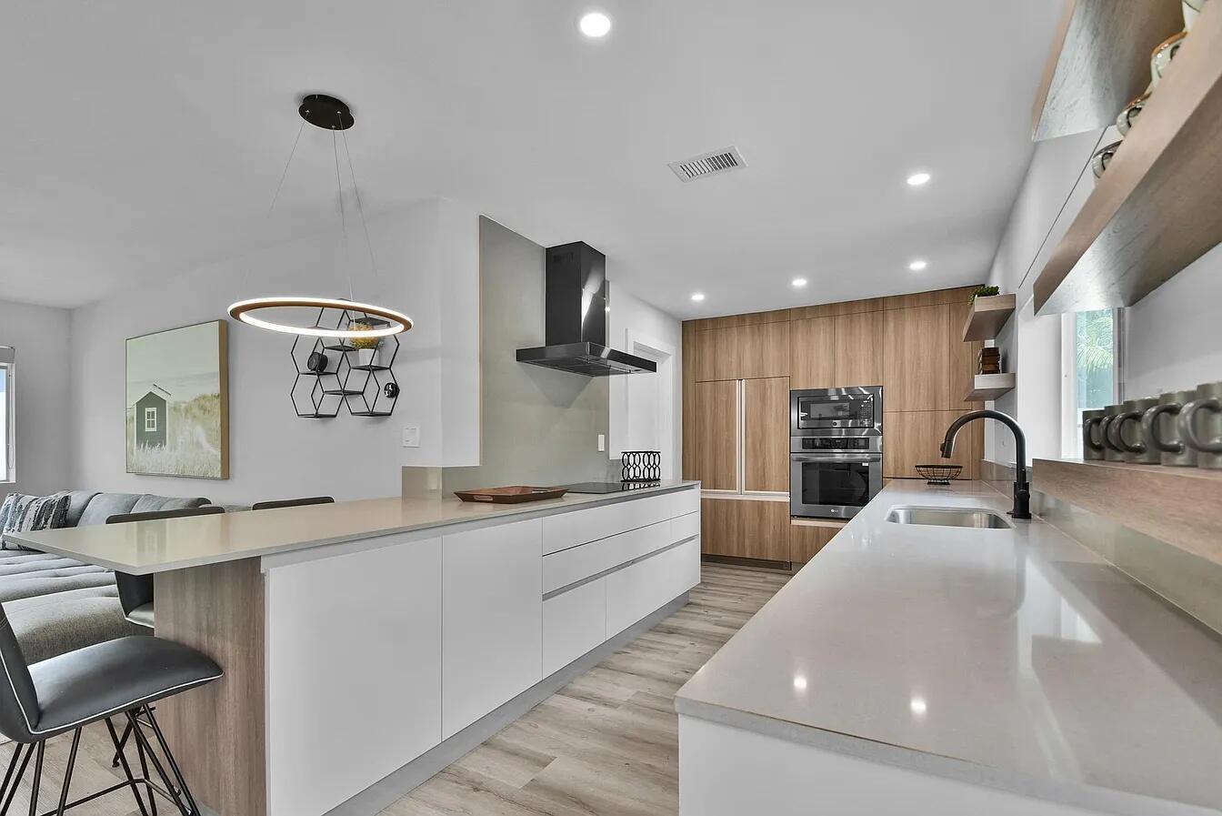 A modern kitchen features white cabinetry, quartz countertops, wood-toned wall panels, and stainless steel appliances — 803 Bluebird Drive, Delray Beach FL 33444, DELRAY BEACH, FL