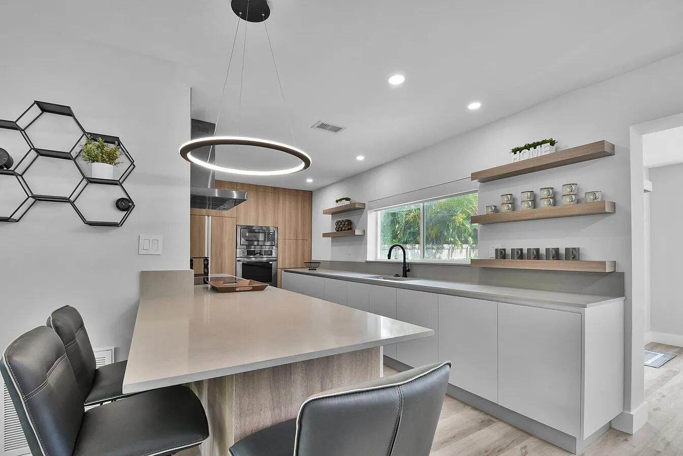 A modern kitchen features a quartz island, light wood cabinetry, open shelving, and gray stools — 803 Bluebird Drive, Delray Beach FL 33444, DELRAY BEACH, FL