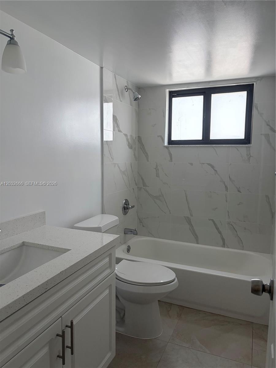 A bathroom features a white vanity, a toilet, and a bathtub with marble-patterned tile walls — 61 W 13th St # 1, Hialeah FL 33010, HIALEAH, FL