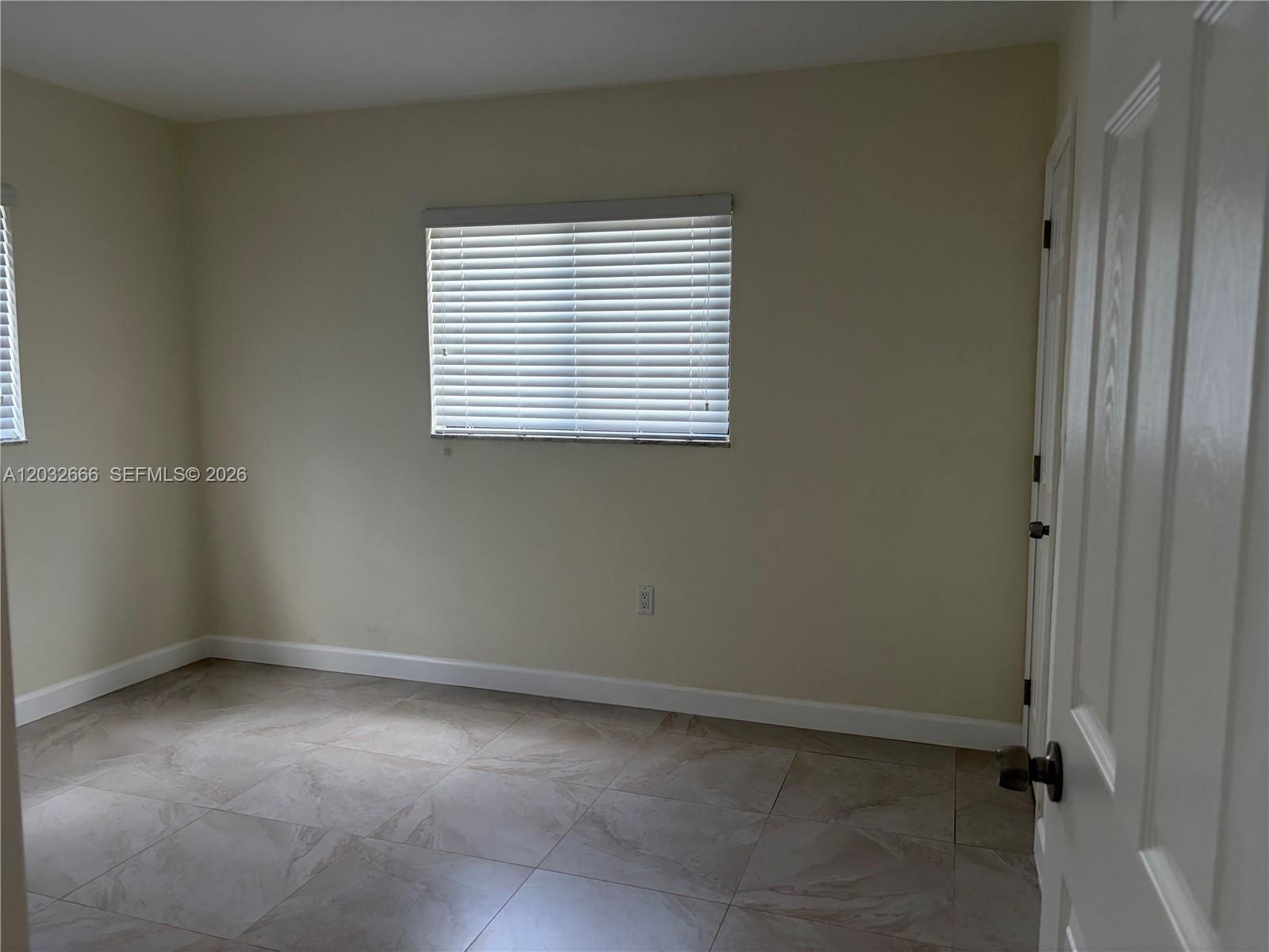 An empty bedroom features light-colored walls, tile flooring, and a window with white blinds — 61 W 13th St # 1, Hialeah FL 33010, HIALEAH, FL