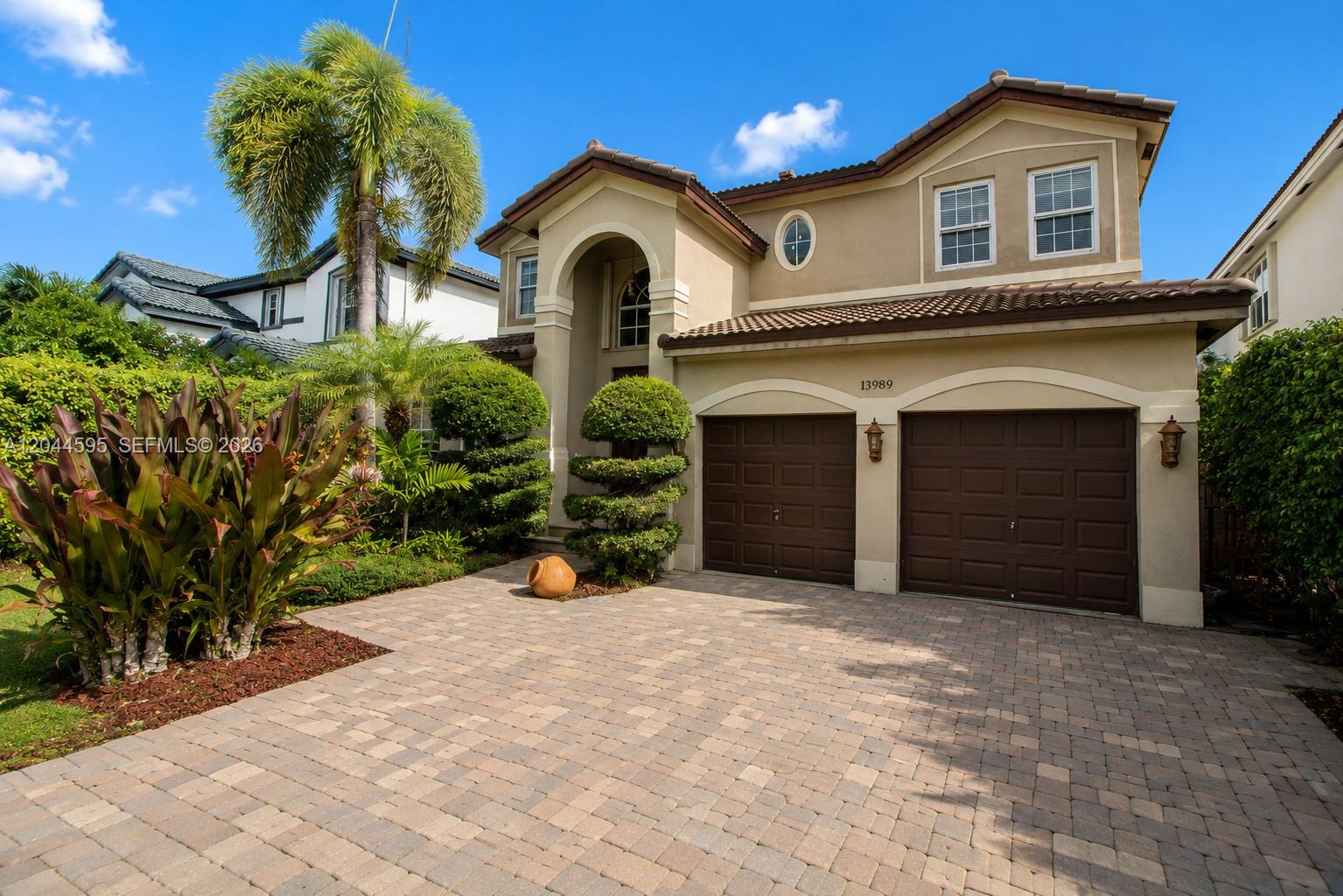 A two-story stucco house features a paver driveway, two garage doors, and lush landscaping — 13989 SW 155th Ter, Miami FL 33177, MIAMI, FL
