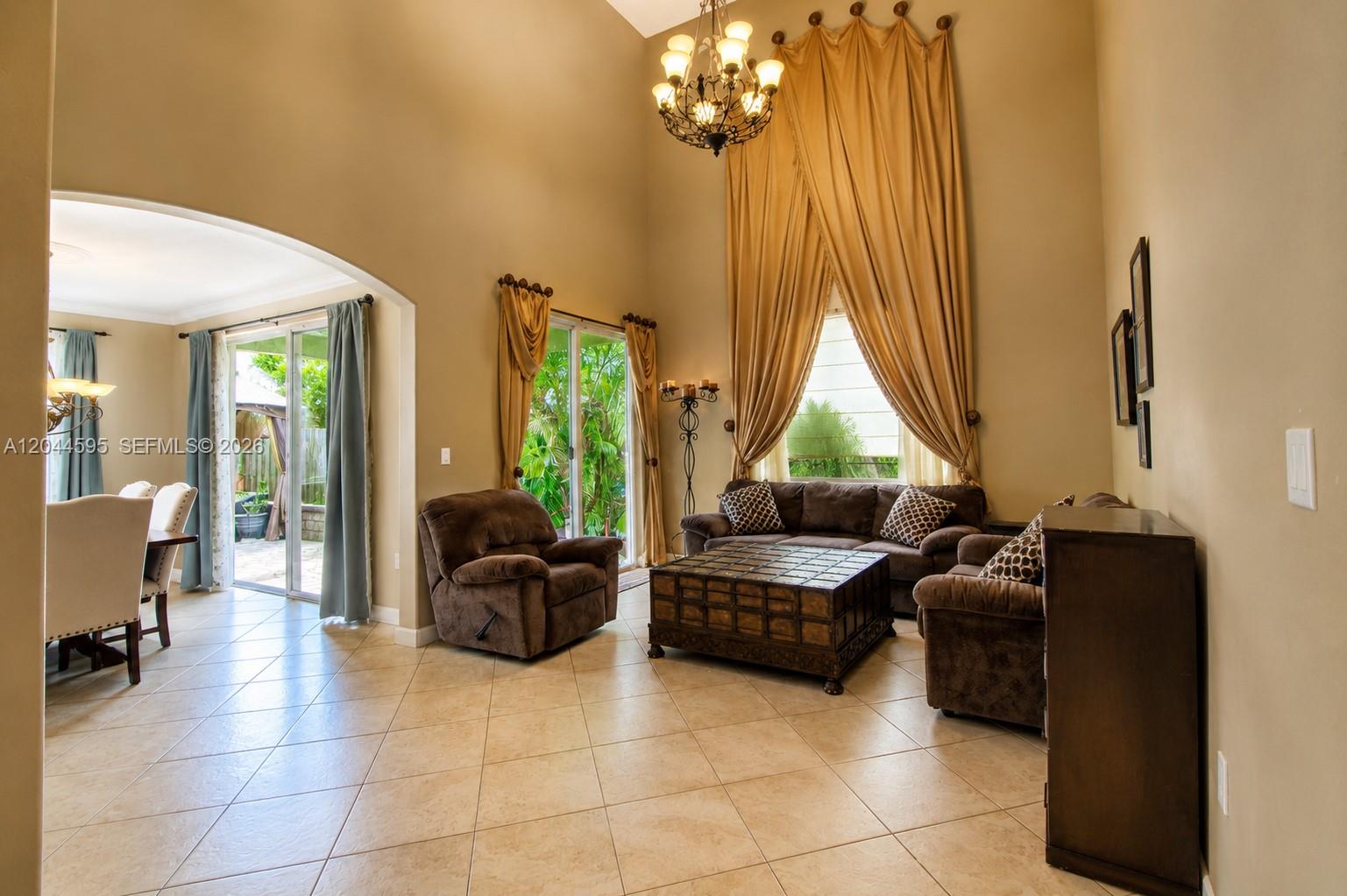 A living room features high ceilings, tile floors, brown furniture, and large draped windows — 13989 SW 155th Ter, Miami FL 33177, MIAMI, FL