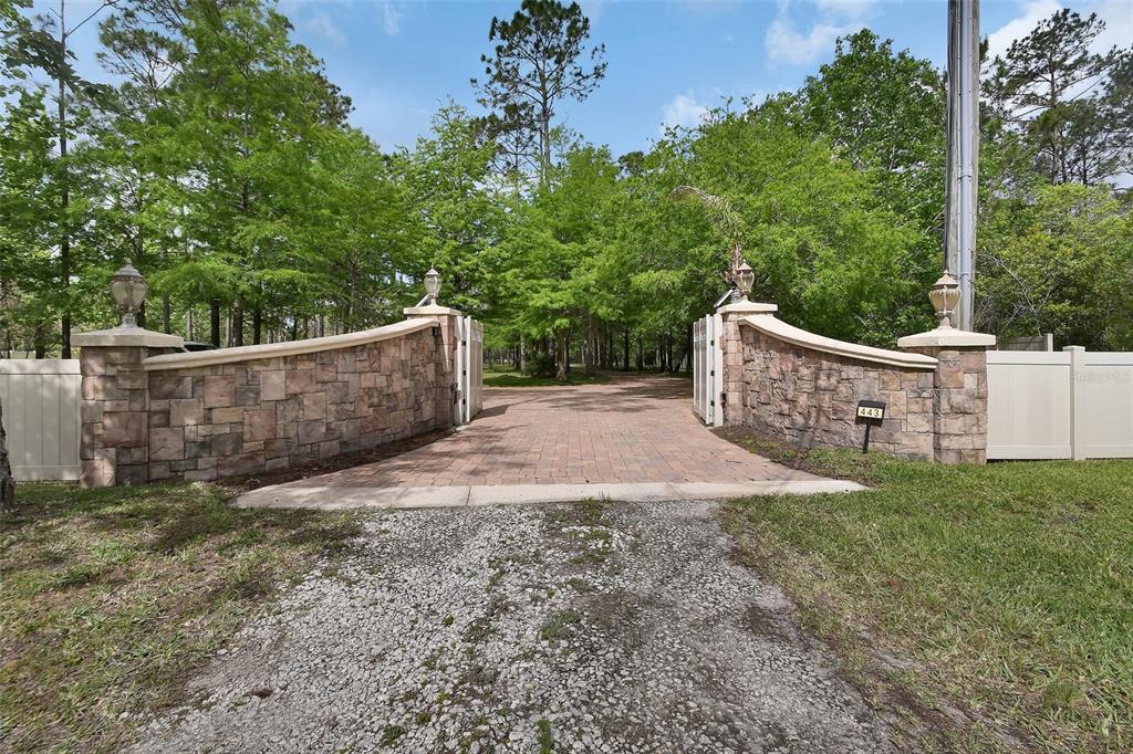 A stone-walled gated entrance leads to a paved driveway surrounded by trees — 443 JUNIPER LANE, ORMOND BEACH, FL