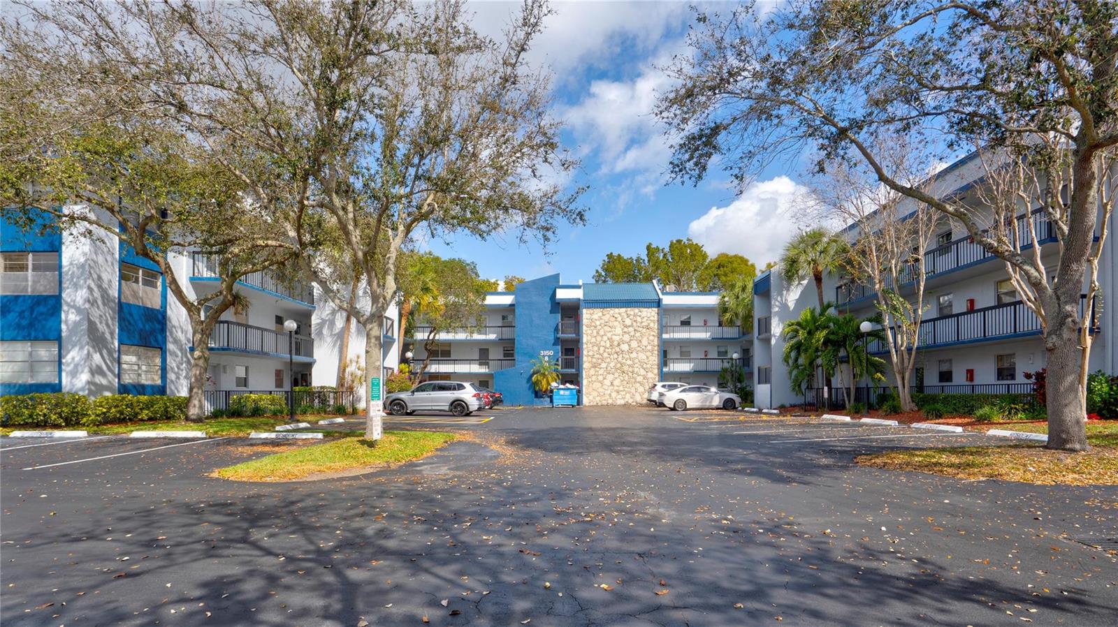 An exterior view of a multi-story apartment complex with a parking lot and trees — 3150 Holiday Springs Boulevard # 311, Margate FL 33063, MARGATE, FL