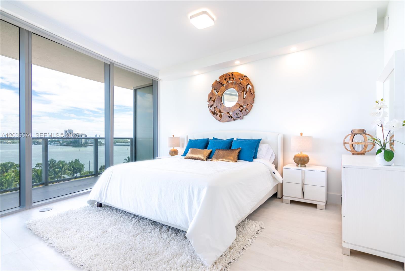 A bright bedroom features a white bed, blue pillows, a round wooden mirror, and balcony views — 488 NE 18th St # 615, Miami FL 33132, MIAMI, FL