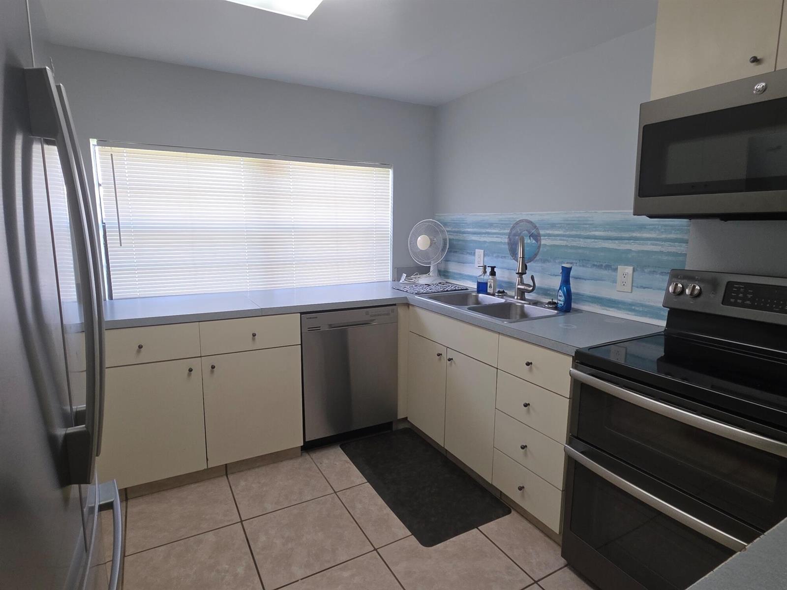 A kitchen features light-colored cabinets, stainless steel appliances, a window with blinds, and tiled flooring — 1019 NW 2nd Street, Okeechobee FL 34972, OKEECHOBEE, FL
