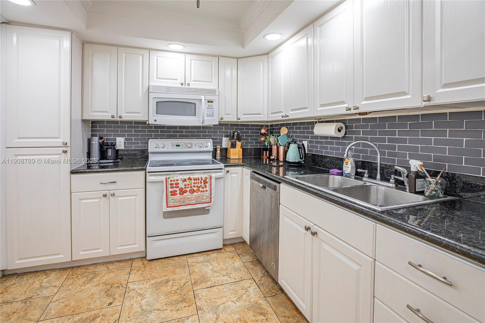 A kitchen features white cabinets, dark granite countertops, a gray subway tile backsplash, and tile flooring — 437 Golden Isles Dr # C, Hallandale Beach FL 33009, HALLANDALE BEACH, FL
