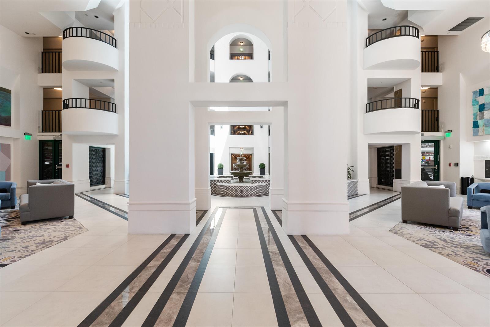 A bright hotel lobby features white walls, arched balconies, a central fountain, and tiled floors — 120 SE 5th Avenue E # 127, Boca Raton FL 33432, BOCA RATON, FL