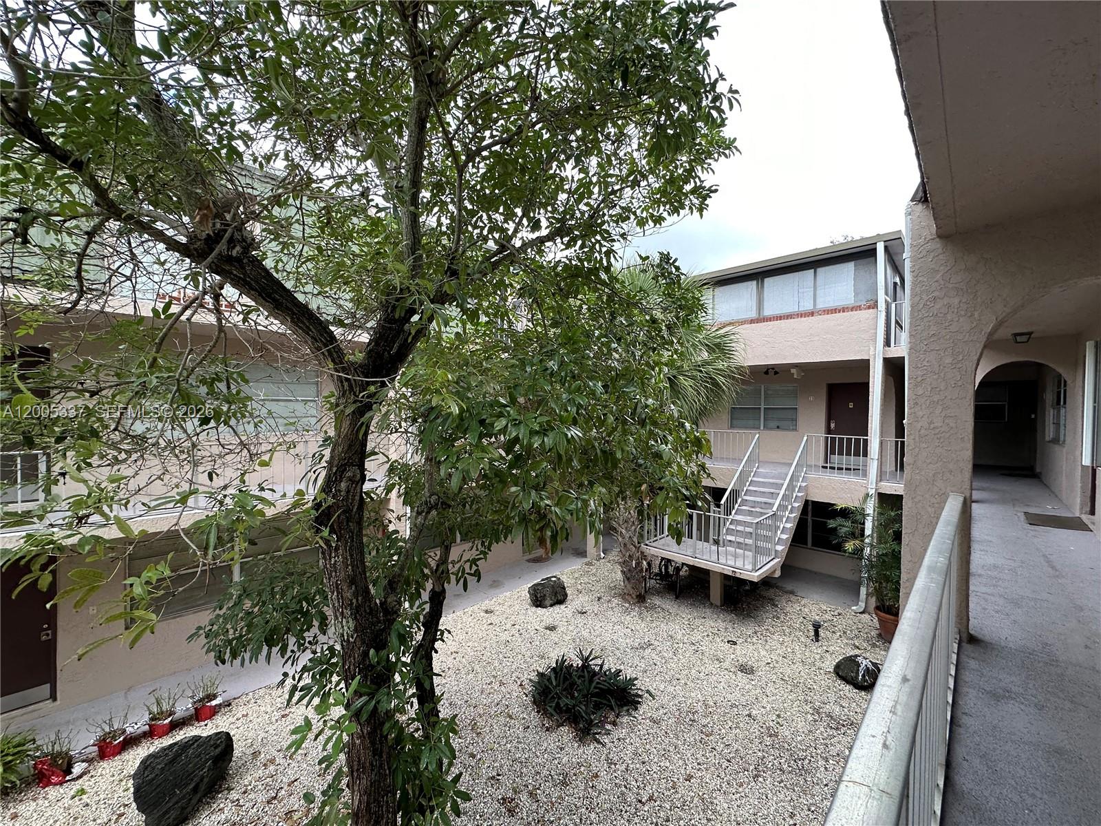 An exterior view of a multi-story apartment complex courtyard with a central tree and staircase — 1100 SE 4th Ave # 32, Deerfield Beach FL 33441, DEERFIELD BEACH, FL