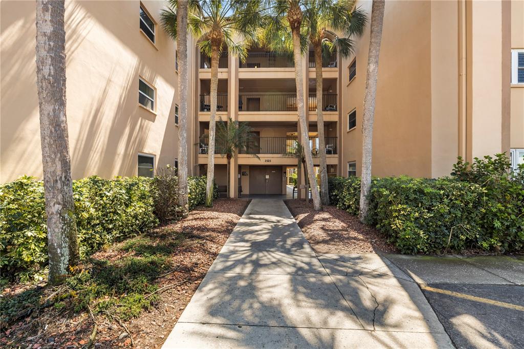 A concrete walkway leads to the entrance of a multi-story apartment building with palm trees — 2525 W BAY DRIVE UNIT C21, BELLEAIR BLUFFS, FL