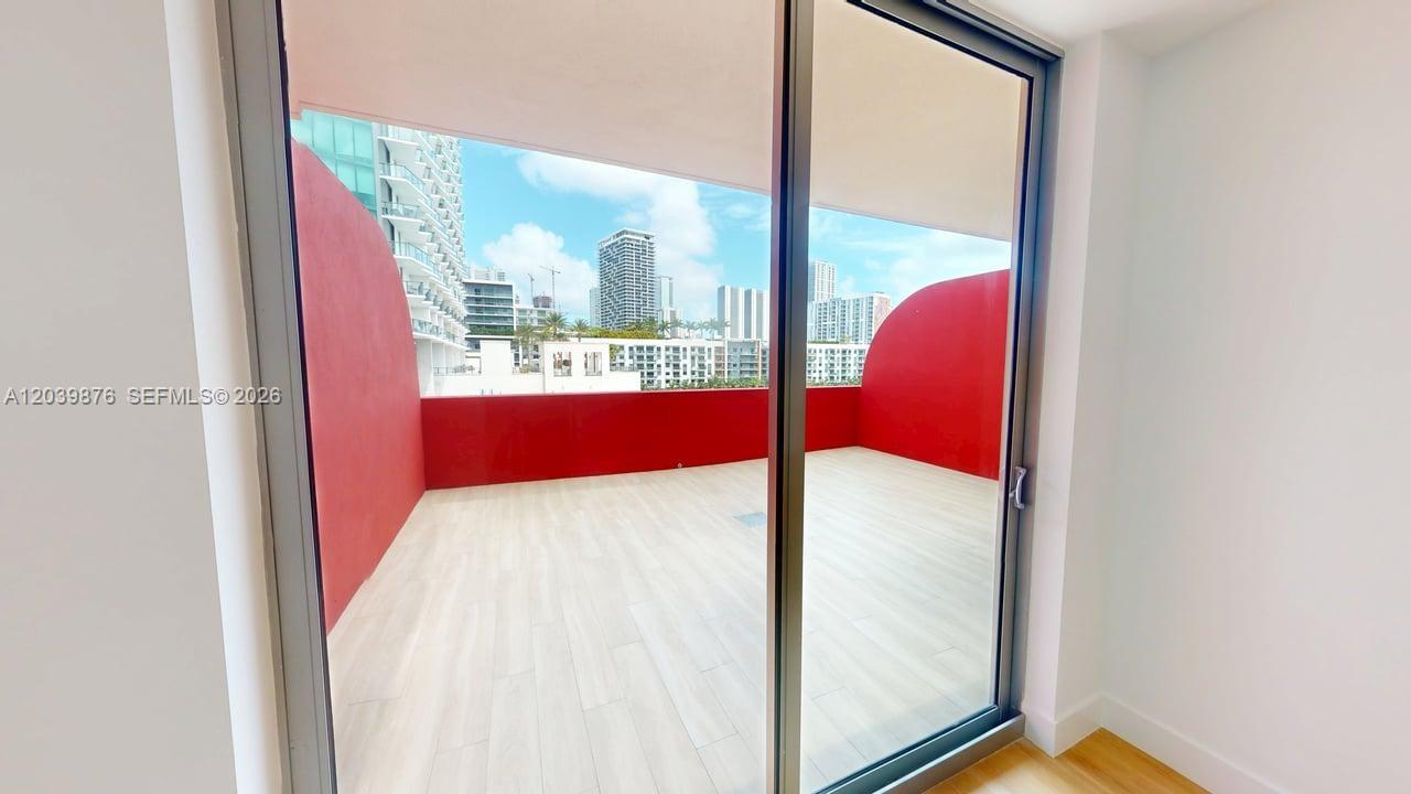 A balcony with red walls and light-colored flooring is viewed through a sliding glass door — 90 NE 32nd Street # 603, Miami FL 33127, MIAMI, FL