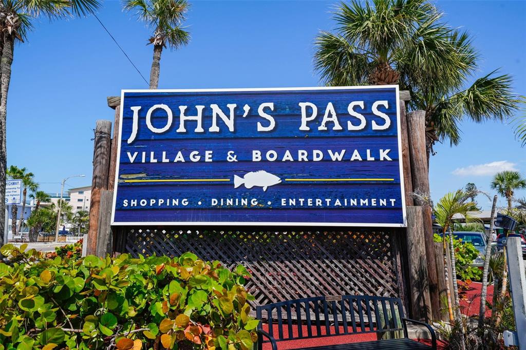 A blue sign for John's Pass Village & Boardwalk stands behind a black metal bench — 13062 GULF BOULEVARD, MADEIRA BEACH, FL
