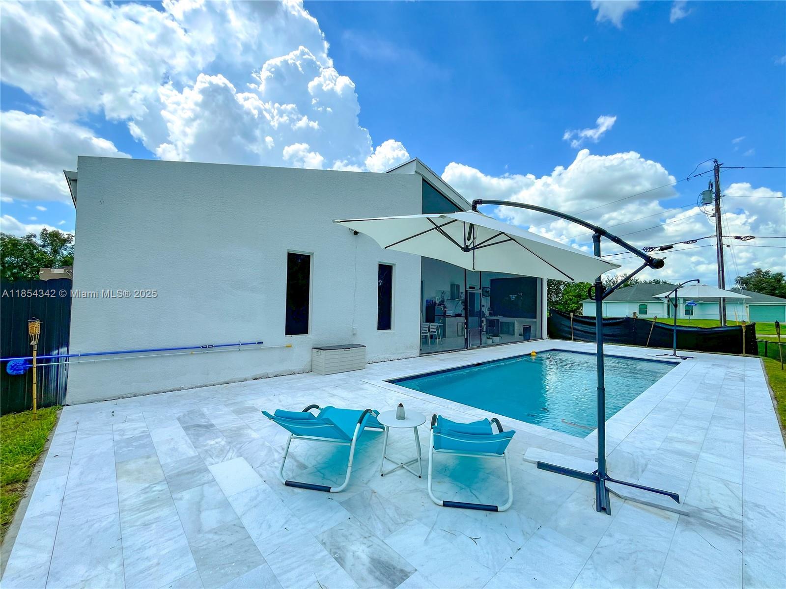 A backyard features a swimming pool, white patio tiles, two lounge chairs, and an umbrella — 395 Pace Avenue, Lehigh Acres FL 33974, LEHIGH ACRES, FL