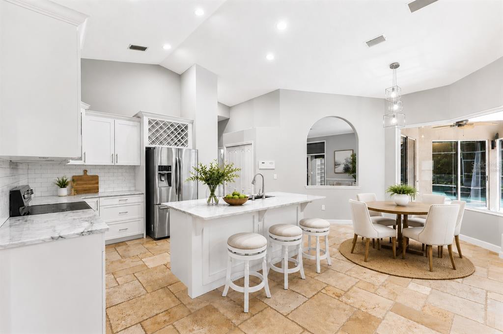 A bright kitchen features white cabinetry, a marble-topped island with bar stools, and stone flooring — 695 SALT LAKE DRIVE, TARPON SPRINGS, FL