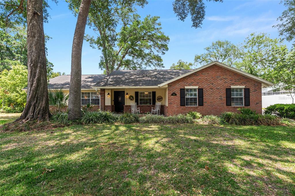 A single-story brick house with a covered front porch sits on a grassy, tree-filled lot — 986 SE 57TH AVENUE, OCALA, FL
