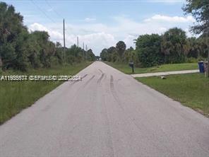 A paved road stretches into the distance, flanked by grassy shoulders and dense green trees — 3215 52nd St W, Lehigh Acres FL 33971, LEHIGH ACRES, FL