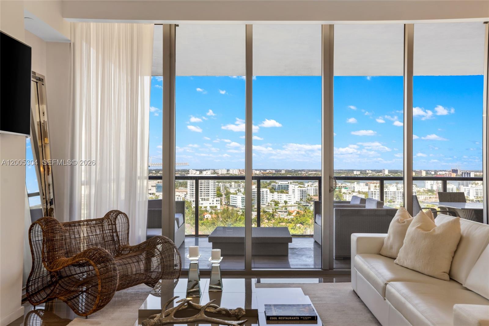 A living room features a wicker chair, white sofa, and floor-to-ceiling windows overlooking a city — 9705 Collins Ave # 1805N, Bal Harbour FL 33154, BAL HARBOUR, FL