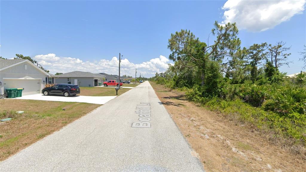 A paved street runs between a residential house on the left and wooded land — 13465 BOATBILL LANE, PORT CHARLOTTE, FL
