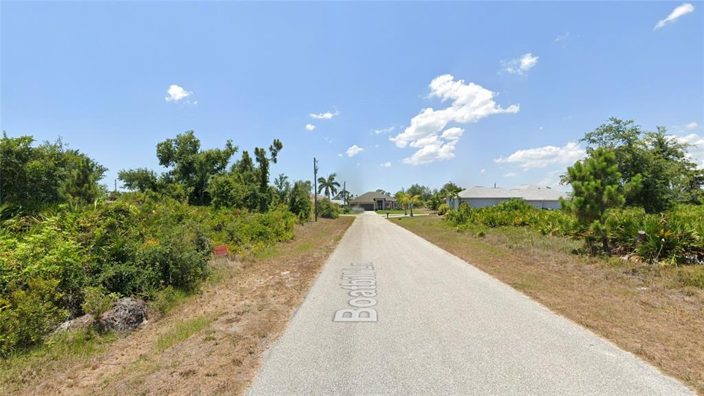 A paved road leads toward a house surrounded by dense vegetation under a blue sky — 13465 BOATBILL LANE, PORT CHARLOTTE, FL