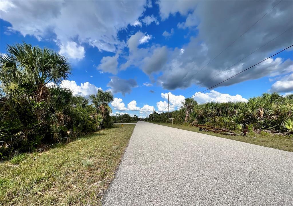 A paved road stretches into the distance, flanked by dense vegetation and palm trees — 14298 MONTMARTE AVENUE, PORT CHARLOTTE, FL