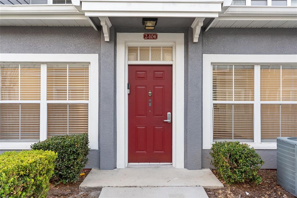 A red front door with a transom window is centered between two windows and bushes — 2608 LODI CIRCLE UNIT 104, KISSIMMEE, FL