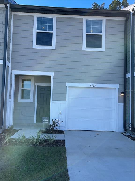 A two-story townhouse exterior features light gray horizontal siding, a white garage door, and a dark door — 6157 DAERR RIDGE UNIT 6157, ZEPHYRHILLS, FL