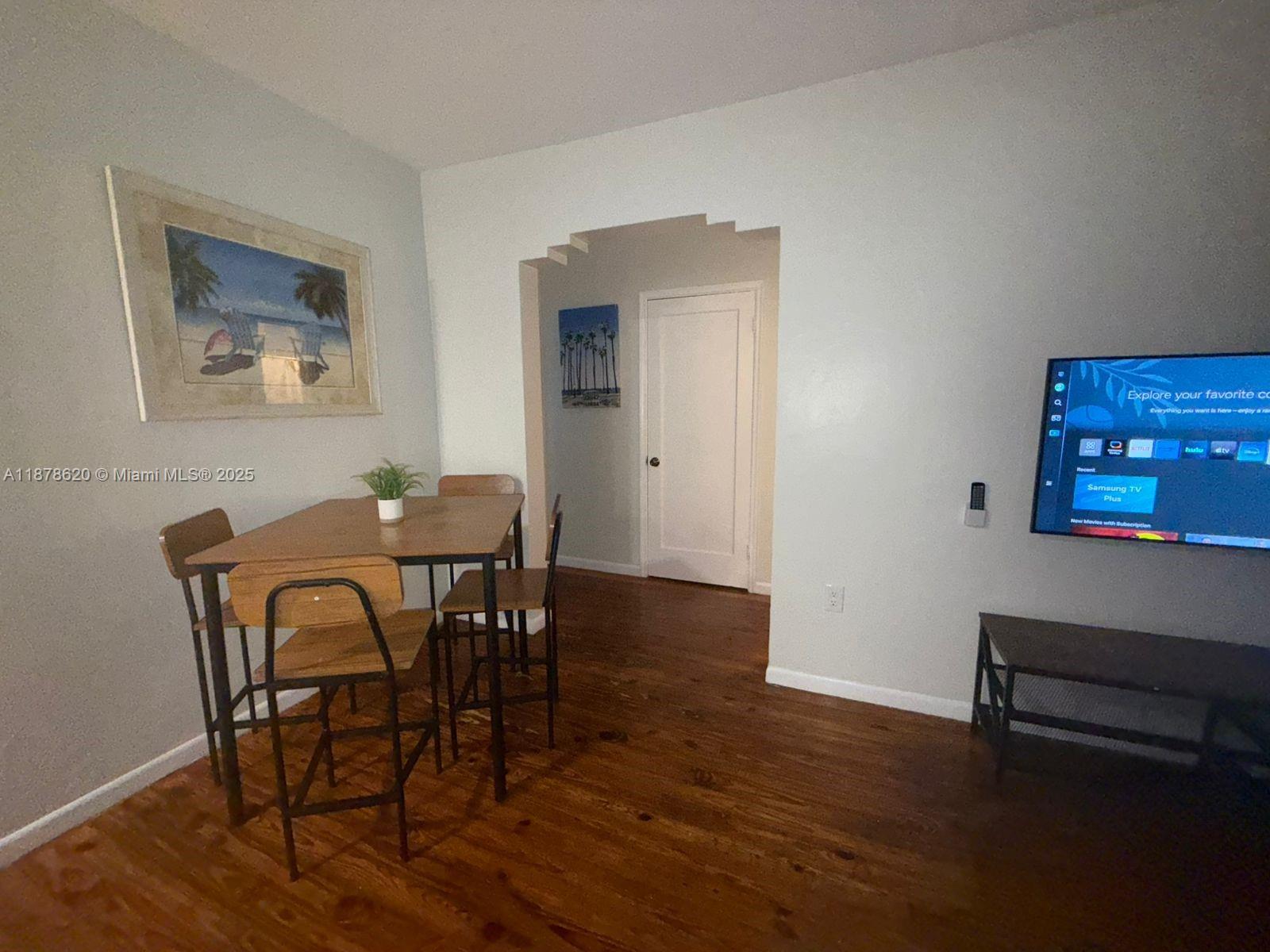 A dining area with a wooden table and chairs sits next to a wall-mounted television — 1289 Marseille Dr # 46, Miami Beach FL 33141, MIAMI BEACH, FL