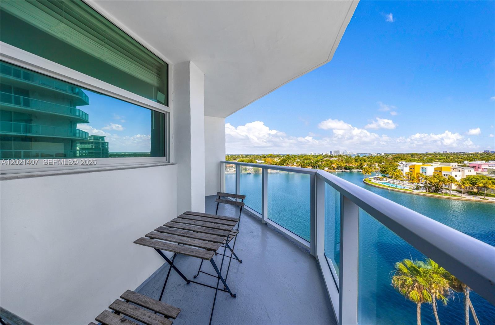 A balcony with a small wooden table and chairs overlooks a bay and city skyline — 5880 Collins Ave # 1007, Miami Beach FL 33140, MIAMI BEACH, FL