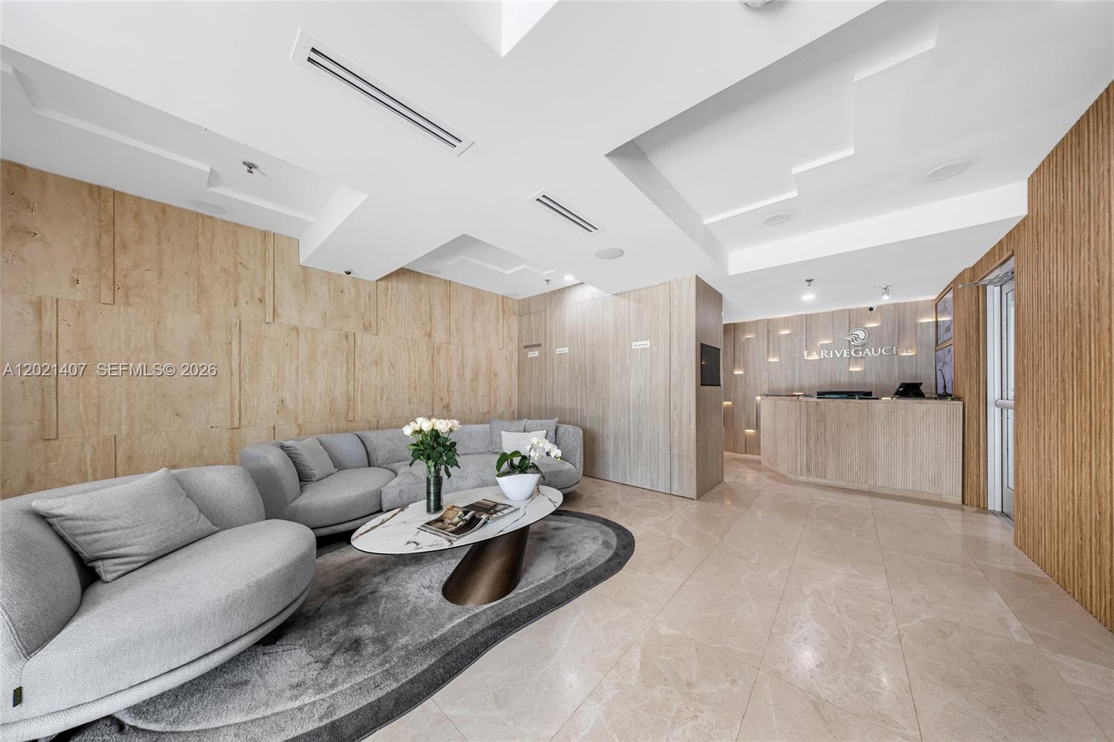 A modern reception area features a curved sofa, marble coffee table, and wood-paneled walls — 5880 Collins Ave # 1007, Miami Beach FL 33140, MIAMI BEACH, FL