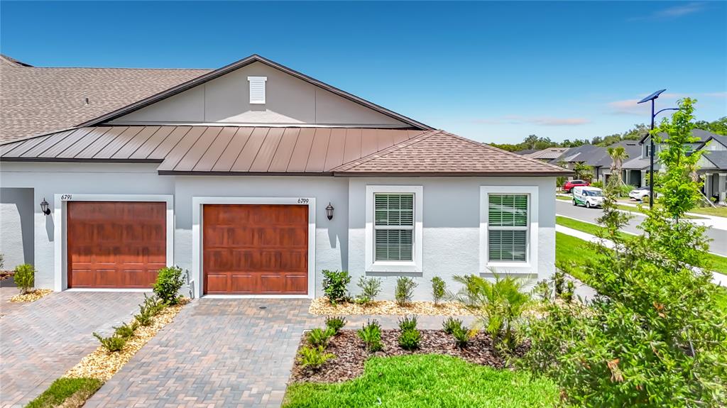 The exterior of a single-story house features two garage doors, light-colored stucco walls, and a metal roof — 6799 HOMEBOUND WAY, NEW PORT RICHEY, FL