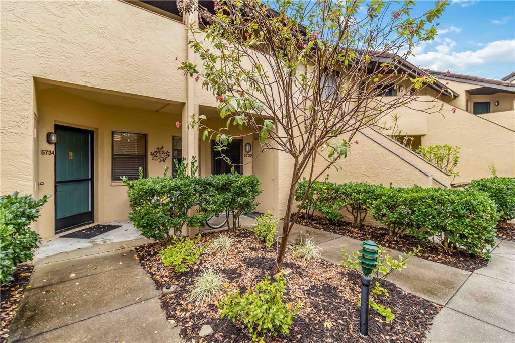 A tan stucco building exterior features two front doors, a small garden, and a tree — 5734 ASHTON LAKE DRIVE UNIT 7, SARASOTA, FL