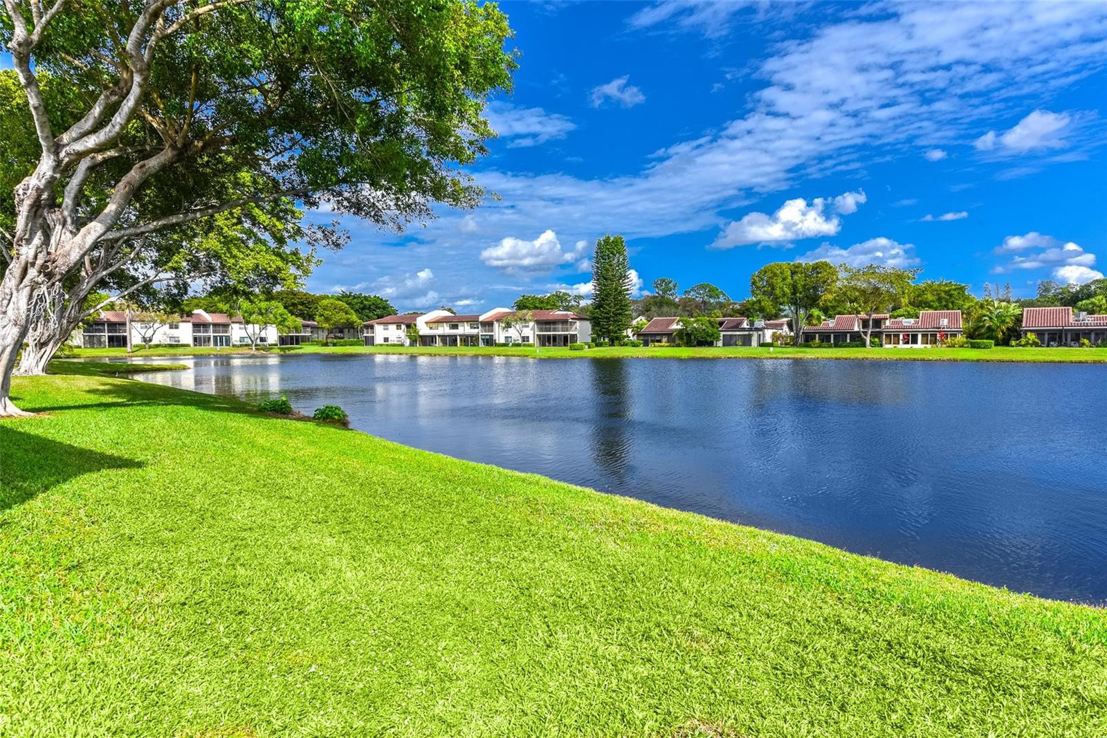 A grassy lawn overlooks a lake with residential buildings in the background under a sky — 21827 Arriba Real # L, Boca Raton FL 33433, BOCA RATON, FL