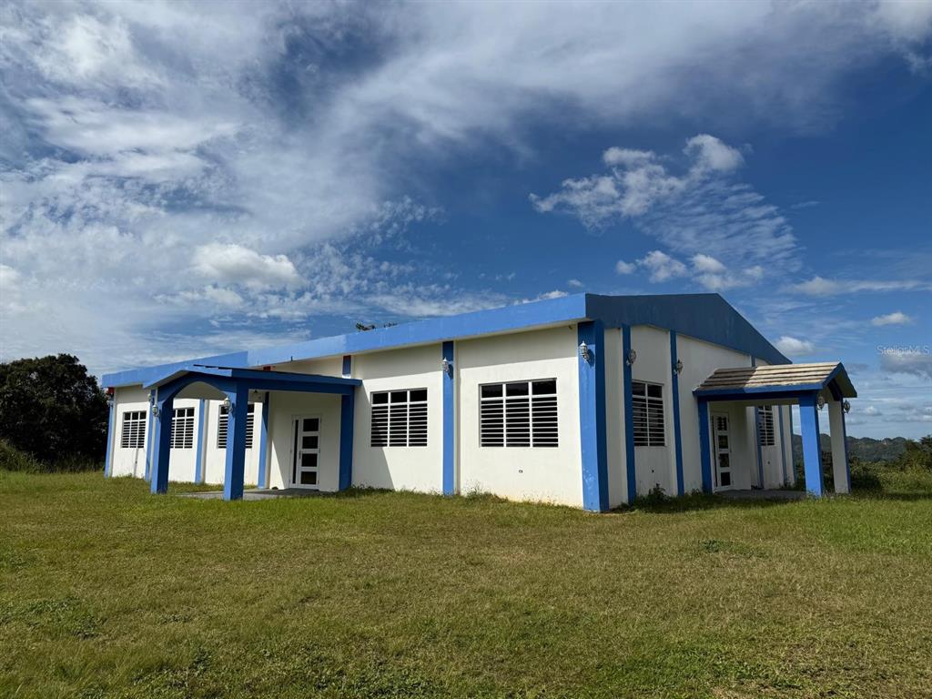 A single-story house with white walls and blue trim sits on a grassy hill — 111 CARR 111 R 602 KM 1.2 INT. BO ANGELES, ANGELES, PR