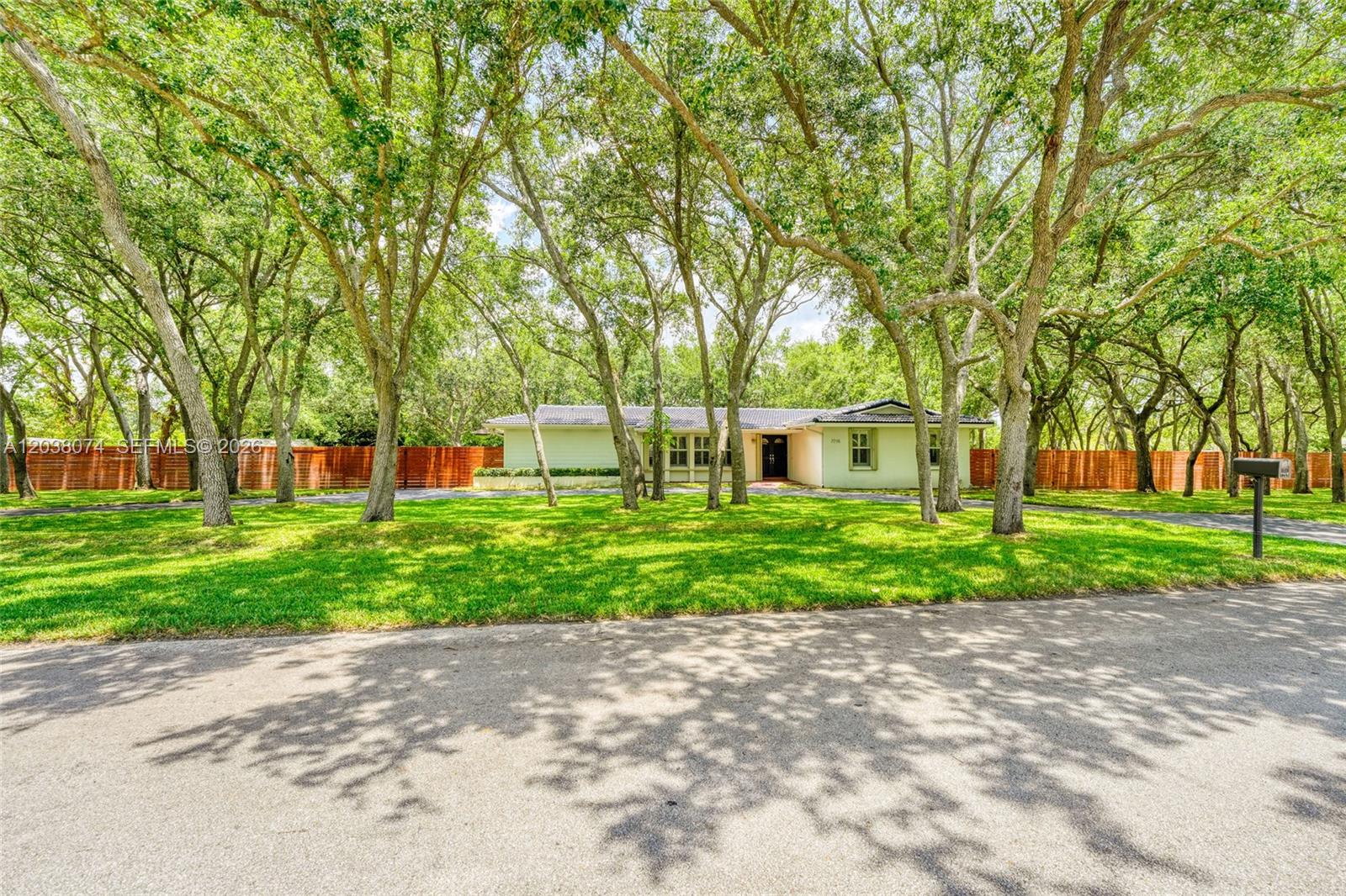 A single-story house with a light-colored exterior sits behind a lawn and many trees — 7715 SW 122nd St, Pinecrest FL 33156, PINECREST, FL