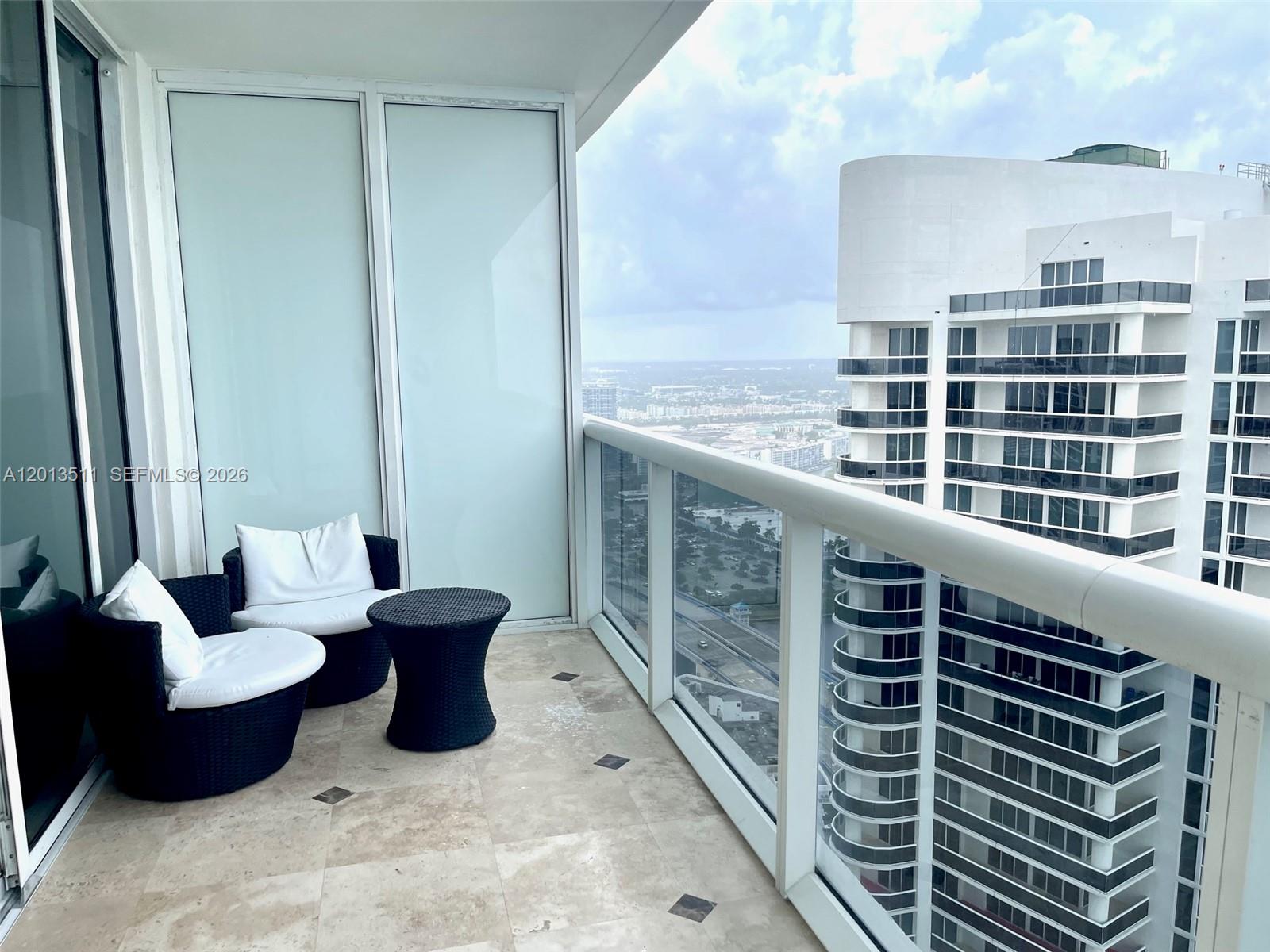 A high-rise balcony features two wicker chairs, a small table, and a glass railing — HALLANDALE BEACH, FL