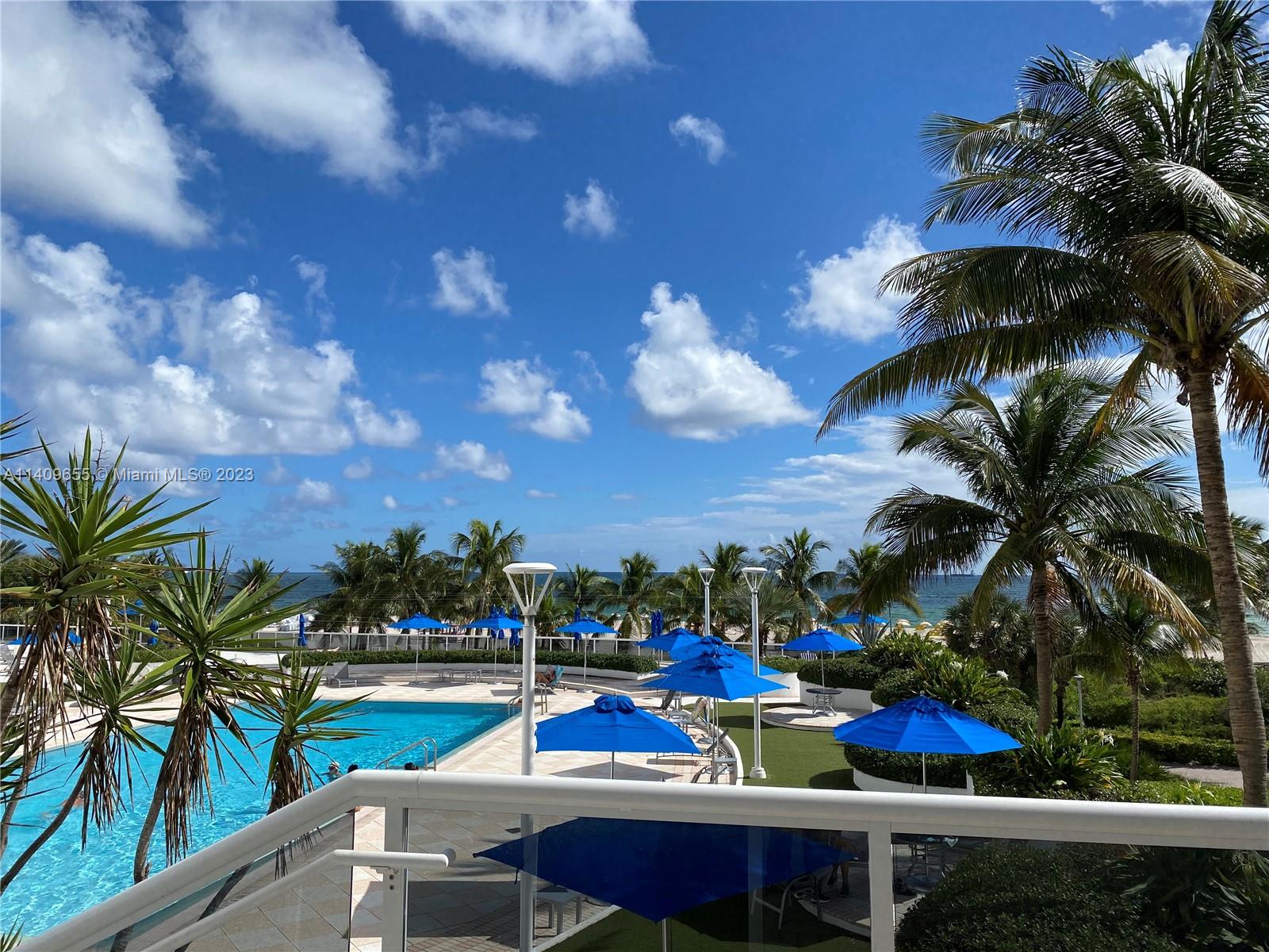 A balcony overlooks a swimming pool, blue umbrellas, palm trees, and the ocean beyond — 100 Lincoln Rd # 939, Miami Beach FL 33139, MIAMI BEACH, FL