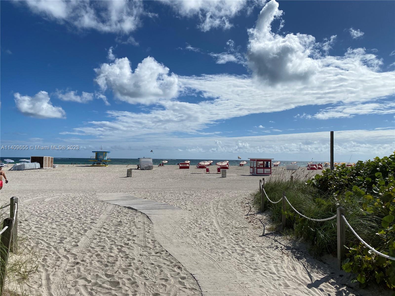 A sandy beach features a wooden walkway, lifeguard stand, and several red beach umbrellas — 100 Lincoln Rd # 939, Miami Beach FL 33139, MIAMI BEACH, FL