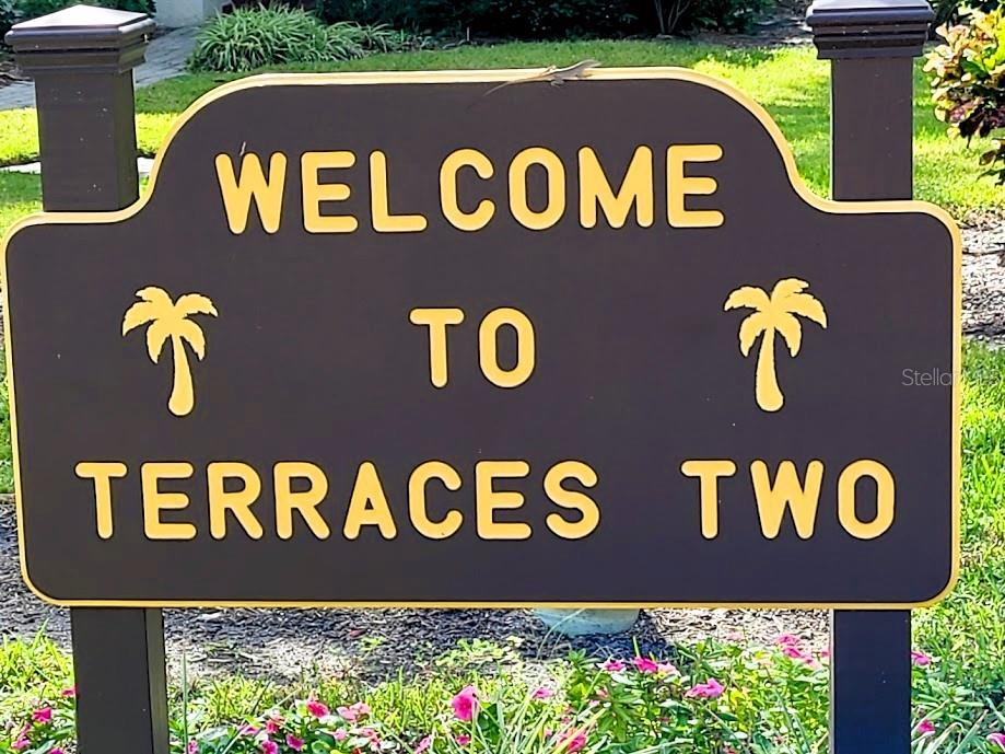 A brown sign with yellow text reads Welcome to Terraces Two, featuring two palm tree icons — 6462 WILD OAK BAY BOULEVARD UNIT 245, BRADENTON, FL